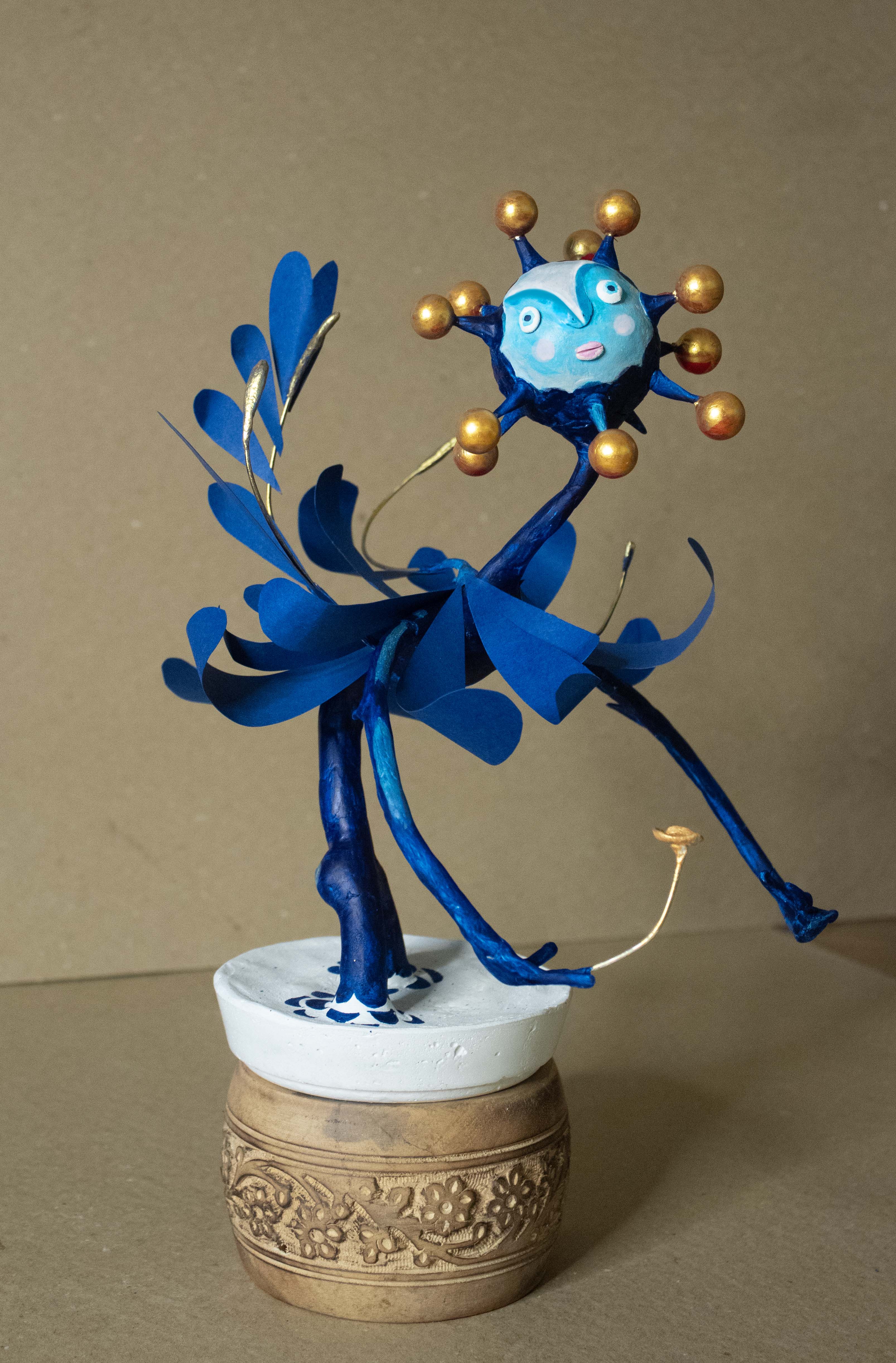 Blue paper mache character sculpture with sun face and gold orbs, dancing on a decorative brass base