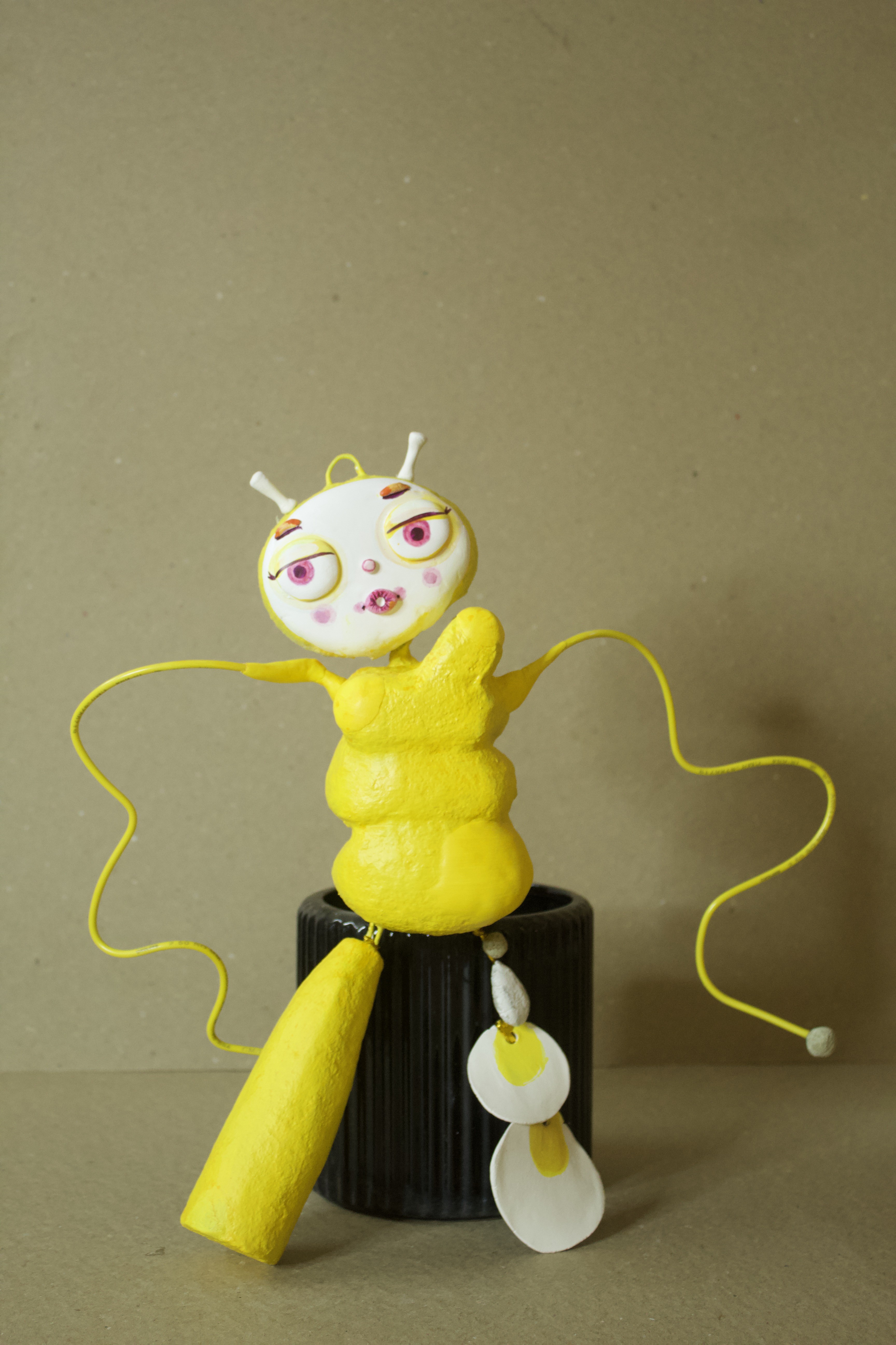 Yellow paper mache character sculpture with white face and pink eyes, seated on a black ribbed pot with trailing yellow wire